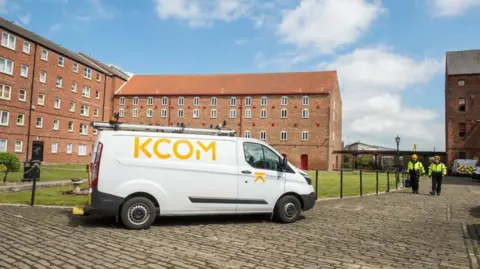 KCOM A white van with the letters KCOM in yellow printed on its right-hand side, which is facing the camera, is parked on a cobbled street. Behind it is a block of flats with small windows. Two people wearing yellow headgear and hi-vis jackets, with dark trousers, walk towards the front of the van.