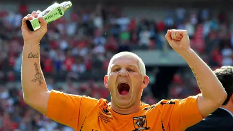 Reuters Dean Windass with arms raised aloft on the pitch at the Wembley play-off final. He holds a trophy in his right hand and is wearing his amber Hull City shirt.