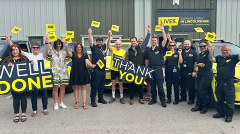 LIVES A group of people, mostly dressed in black, stand front of a line of emergency response vehicles. One is holding up a sign that reads "well done." Another is holding a sign that reads "thank you". The rest have placards. In the centre, a cyclist in yellow holds up an arm in a celebratory pose.