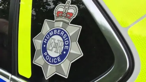 BBC A close-up of a Humberside Police logo on the rear passenger window of a police car. The logo is a silver star with a crown on top with "Humberside Police" written around "E II R".