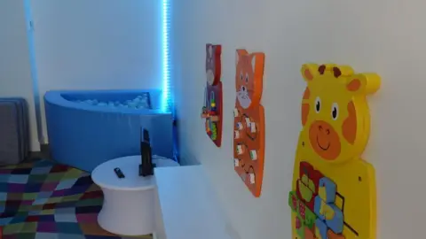 Hull City Council A room with brightly-coloured wall-mounted puzzle games, a ball pool and fluorescent blue light in the background.