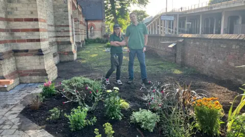 Giroscope Woman wearing green T-shirt, cap and dark jeans with man wearing green T-shirt with blue jeans stand on a patch of soil behind plants, next to a paved area. There is a brick and stone church building to one side of them and a brick wall and another building to the other.