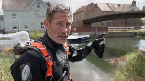 BBC A man with grey hair and stubble wearing a black wet suit, with an oxygen tank on his back points at a bridge above the River Hull. He's wearing black rubber gloves. Behind him is white motor boat and a pub with light green rendered walls.