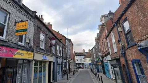 Google A narrow paved town centre street, complete with shop fronts, pavement bollards and other street furniture.