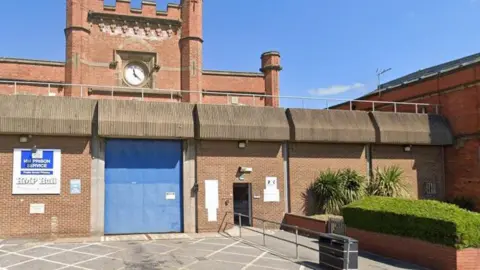 Google The outside of HMP Hull, as denoted by a sign on a wall. A large blue entry door, which is closed, is visible, with high walls on either side. A clock tower protrudes from behind.