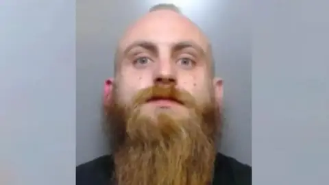 Durham Police Mugshot of a man with a little hair in the middle of his head and a long brown beard staring at the camera. He is wearing a black shirt.
