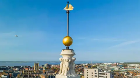 Hull City Council Close up of the time ball on top of Hull's Guildhall it is gold and is mounted on a black pole with a golden sailing ship on the top in the background is the city skyline and the Humber Estuary