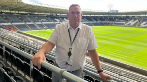 BBC A man with grey hair and wearing a white shirt, grey trousers and black lanyard stands among black seats in a large sports stadium with his hand on a steel rail. A grass pitch can be seen in the background.