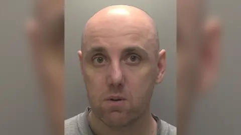 Humberside Police A police head shot of a bald man wearing a grey t-shirt. He is stood in front of a grey wall. 