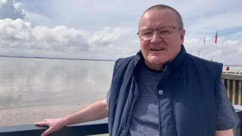 BBC/Sally Fairfax A bald man, with glasses is standing in front of a railing by the river Humber. He is smiling at the camera and wearing a navy blue t shirt and navy blue body warmer.