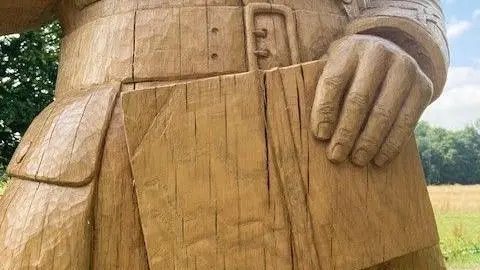 A close up of a wooden statue, showing visible vertical cracks. The statue is a yellow/golden colour and depicts a man holding a book and wearing a rain coat.
