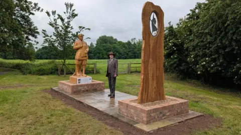 East Riding of Yorkshire Council A man wearing a trilby hat and brown three-piece suit is standing on paving stones in between two wooden sculptures, erected either side of him. One of the sculptures is a man holding a book, while the other has a small circle cut out with a silhouette of a woman dancing inside. The sculptures are in grassland.