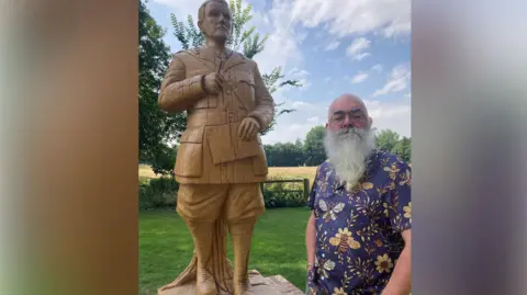 BBC A bearded man in a blue shirt decorated with yellow bees stands next to a wooden statue of JRR Tolkien, that is showing cracks.