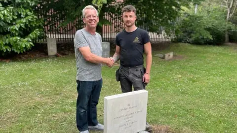 BBC A man with short grey hair wearing a grey patterned t-shirt and blue jeans stood next to a gravestone in a graveyard. He is shaking hands with a man with short brown hair who is wearing a black t-shirt and black trousers. The grass is green and green trees can be seen in the background.