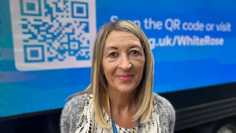 BBC News A woman with long blond hair and wearing a grey jumpers smiles as she stands in front of a blue van. On the side of the van is displayed a large white QR code and text (partially obscured) reading, "Scan the QR code or visit ycr.org.uk/whiterose".