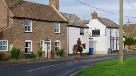 Paul Harrop/Geograph/Creative Commons A rider on a brown horse is seen on a road in the centre of the image in front of a row of old brick and white-washed houses, including a pub, with looks to date from the 18th or 19th Centuries. To the right, a telegraph pole stands on a green grass verge.