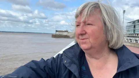 BBC / Gill Sennett A woman with light-grey hair in a bob wears a blue jacket and stands on a promenade staring across the grey-brown waters of a wide river estuary. White railings and a large glass building can be seen to her right. The sky is blue and dotted with white clouds.
