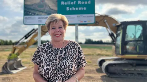 Becki Bowden/BBC A woman with short blonde hair wears a leopard-print top and long necklace. She is smiling as she stands in a field in front of a large sign reading "relief road scheme". A large yellow excavator is parked behind the sign.