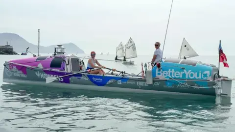Seas the Day Two female rowers in their 20s on a grey rowing boat in a blue sea. The boat has blue branding on the right end and pink branding on the left. One of the women, Miriam Payne, is sitting down with oars in her hands. She is wearing an orange cap, sunglasses and white vest top. Jess Rowe is standing up, wearing sunglasses, blonde hair tied back and a white t-shirt. There are three small boats with white sails in the background.