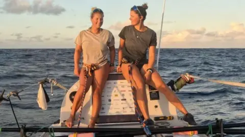Seas The Day Jess Rowe and Miriam Payne are perching on their small boat which is at sea under a light-blue sky in low light. Jess wears a white T-shirt, while Miriam wears a black T-shirt.