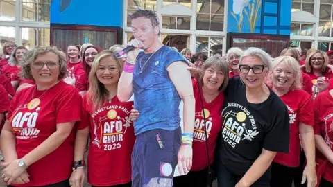 BBC Female members of the All For One Choir stood in a large group inside Paragon Station. All of them are wearing red t-shirts with the words "Disneyland Paris: AFO Choir 2025" on them in white. One woman on the front row is wearing a black t-shirt. The are posing with a cardboard cut out of Chris Martin. 