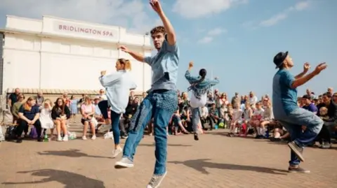 East Riding of Yorkshire Council Four street performers, made up of two men and two women wearing denim jeans and denim trousers, are performing a dance on the street outside a theatre with people sat down watching them.