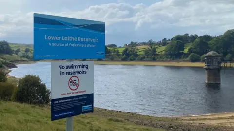 BBC/Grace Wood A blue sign reads: Lower Laithe Reservoir. Below it a white sign reads: No swimming in reservoirs. Behind the signs is a low-level reservoir.