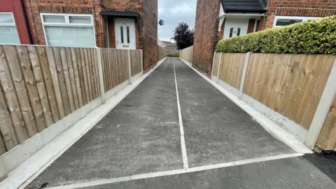 A newly resurfaced tarmac alleyway stretches into the distance between wooden fences and two red brick houses.