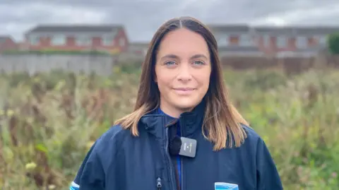 BBC A young woman with blue eyes and shoulder-length light brown hair smiles at the camera. She is wearing a blue waterproof jacket and standing in front of an area of wild flowers and grasses with modern housing visible behind a wooden fence in the background.