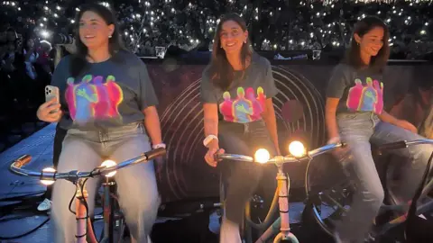 Coldplay Three girls wearing blue jeans and grey Coldplay t-shirts are sitting on bikes and peddling to generate electricity at a Coldplay concert in Amsterdam. They all have shoulder-length dark hair and there are thousands of people in the background with their torches on mobile phones lighting up the sky.