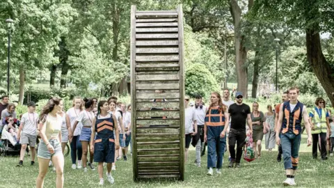 Autin Dance Theatre People of various ages walk with a large wooden wheel structure through green parkland, accompanied by three people wearing navy blue outfits with orange trim, and one person wearing a yellow hi-vis jacket.
