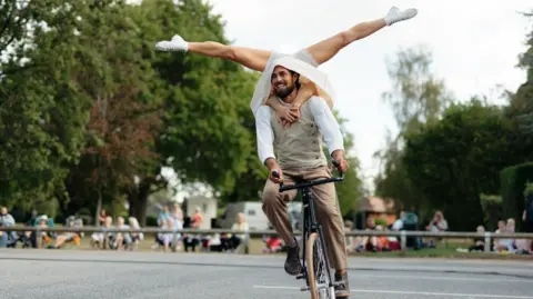 Tom Arran A man rides a black cycle with only one wheel visible, while a second performer balances upside down on his back, arms wrapped around the man's shoulders, legs in straddle splits in the air. The upside-down performer wears white shorts, boots, and what appears to be a skirt, which is draped around the cyclist's shoulders. The cyclist has dark hair and facial hair, and is wearing light brown trousers and shoes, a beige sleeveless jumper and a white shirt.