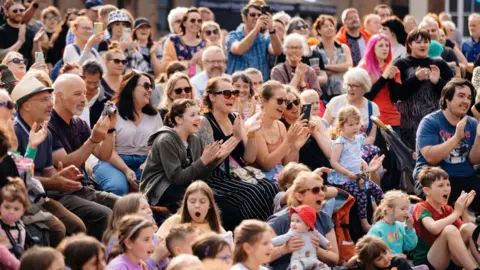 Tom Arran A large crowd of women, men and children applauds. Some are holding babies. They are mostly seated outside, on a bright and sunny day, with some people standing to the rear of the crowd. Some people are smiling, others are open-mouthed, some appear to be recording what they see on their phones and cameras.