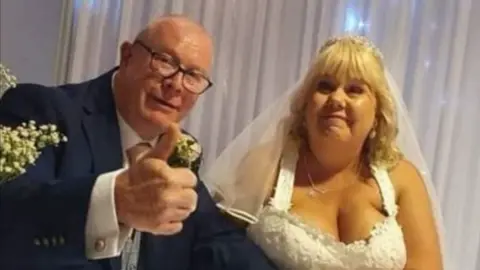 Family handout A woman in a white wedding gown with her husband, who is wearing a navy suit, on their wedding day. She has shoulder-length curly blonde hair while he has short white hair and is wearing glasses. He has a thumb up to the screen.