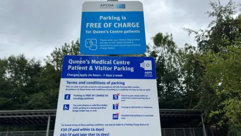 A parking sign. There are two boards, one above the other. The top board says that parking is free of charge for Queen's Centre Patients, with details about entering your car registration on a tablet in the centre reception. The board below warns that ANPR is in use, with terms and conditions of parking. It warns that a breach of the conditions could lead to a charge of £30 if paid within 14 days, rising to £60 if paid later than that.