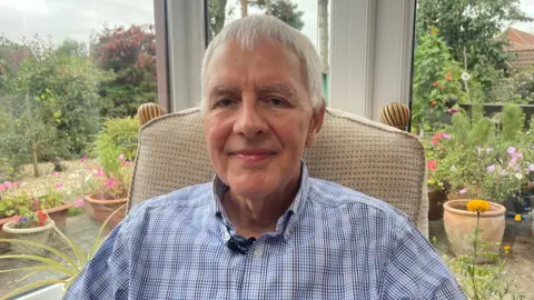 BBC A man in his 60s with short white hair, looking towards the camera. He is wearing a light blue check shirt. He is sitting in a beige chair in his conservatory, with a view of his garden behind him. There are lots of plants and flowers on show in the garden.