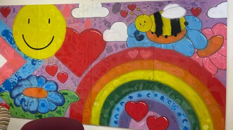 BBC News A colourful painted mural includes red love hearts, a big yellow smiley face, clouds, a rainbow, flowers and a black and yellow bee, along with a series of handprints.