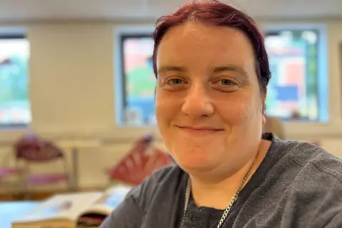 BBC News A close-up of a smiling woman who has tied-back maroon-coloured hair. She is wearing a grey T-shirt and silver necklace and sitting in a community space within a modern church hall.