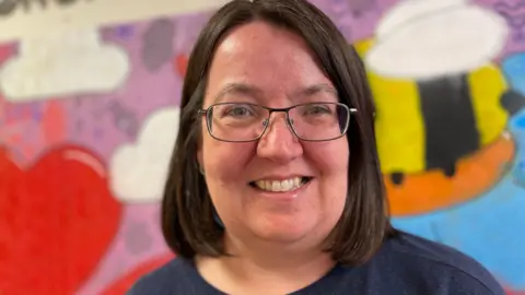 BBC News A close-up of a woman with her hair in a brown bob. She is smiling and wearing glasses and a navy blue top. She is standing in front of a colourful mural, which includes red love hearts and a black and yellow bee. 