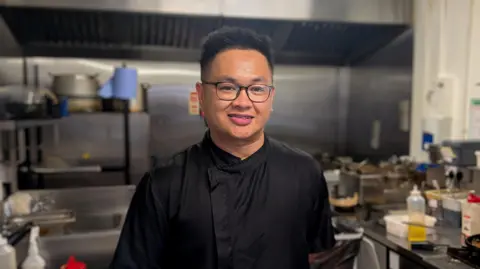 BBC / Josh Gorroño Chapman Ryan Albano smiles into camera in a black chef's uniform. He has dark hair and black glasses and is standing in a commercial kitchen surrounded by cooking utensils and ingredients.