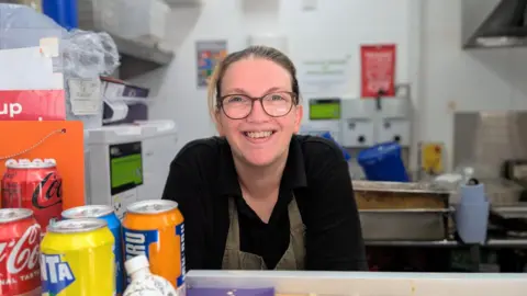BBC / Josh Gorroño Chapman Rachel Stainton stands behind a counter in a food service area with drinks cans and other items on display. She is smiling, has blonde hair and wears glasses. Kitchen equipment and wall signs are visible in the background.