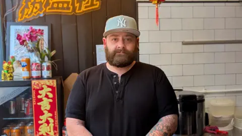 BBC / Josh Gorroño Chapman John Leedham looks into camera. He wears a black shirt and Yankees cap and stands in a kitchen area with containers, bottles, and utensils on the counter. A fridge with jars and flowers sits beside a neon sign and a red lantern hangs from the ceiling.