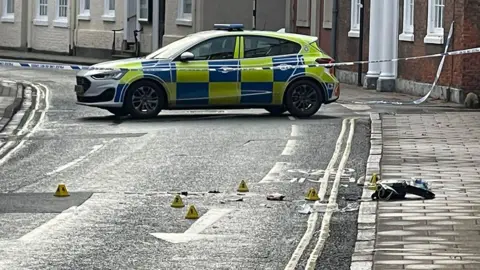 A view of the scene in Beverley with a police car and cordon tape and evidence markers on the road and the pavement.
