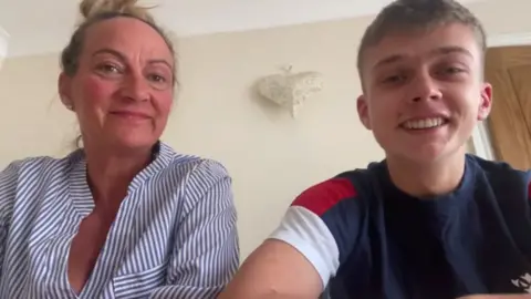 A young man sits next to his mother, They are both smiling at the camera. They are sitting in a room in a house, with a piece of art on the wall behind them, and a door is closed behind them. This is a screengrab from a video call, and they are both looking slightly down at the camera.