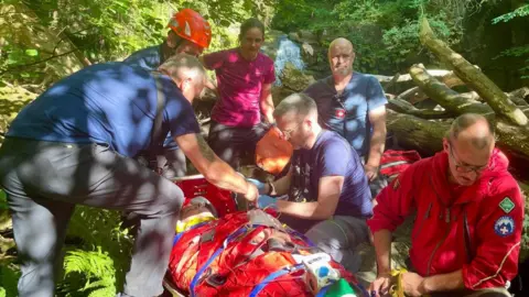 SRMRT Five people surround another person strapped to a stretcher. One is wearing a helmet, another is wearing a jacket with a mountain rescue logo on it. They are outdoors in an area full of greenery and fallen trees.