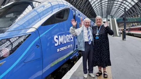 Lumo and Hull Trains A man with short white hair, a white shirt, a blue patterned tie and dark trousers standing with a woman who has light shoulder-length hair, a blue patterned dress, a lanyard and sandals, next to a blue train with "Smurfs" branding on it. They are standing on a platform in a railway station and waving at the camera.