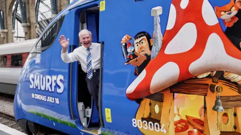 Lumo and Hull Trains A man with short white hair, a white shirt, a blue patterned tie and dark trousers, waving from the door of a blue train with "Smurfs" branding on it. There is a LNER Azuma train in the background within a brick and steel railway station.
