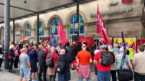 BBC A group of people gathering around a man speaking with a microphone outside of Hull train station. Some are holding flags.