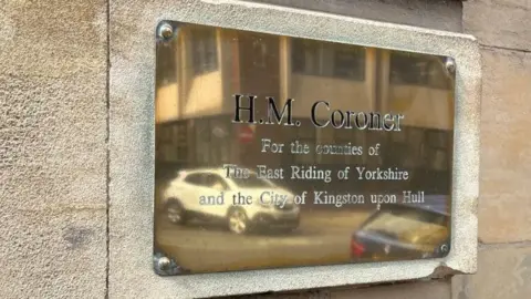 A gold plaque on a white and sandy wall that reads "H.M. Coroner for the counties of The East Riding of Yorkshire and the City of Kingston upon Hull".