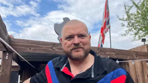A man - Alex Smith - stood by a wooden structure outside with a union jack flag erected behind him. He is bald with brown facial hair and is wearing a black polo shirt with blue and red stripes on. 